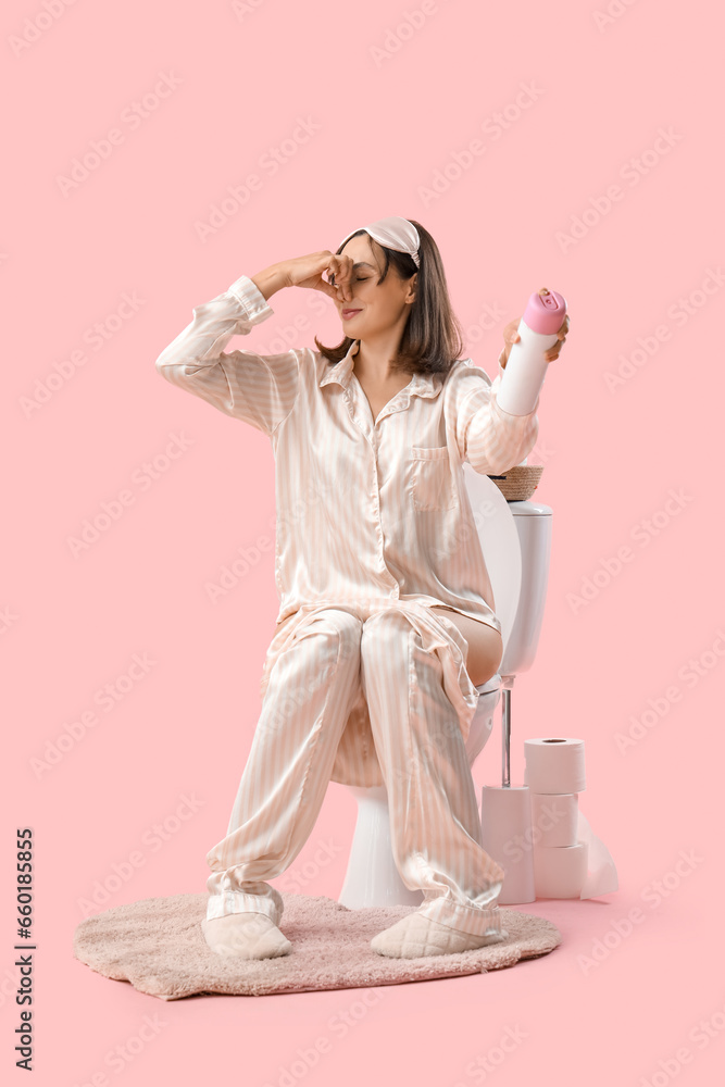 Displeased young woman in pajamas with air freshener sitting on toilet bowl against pink background