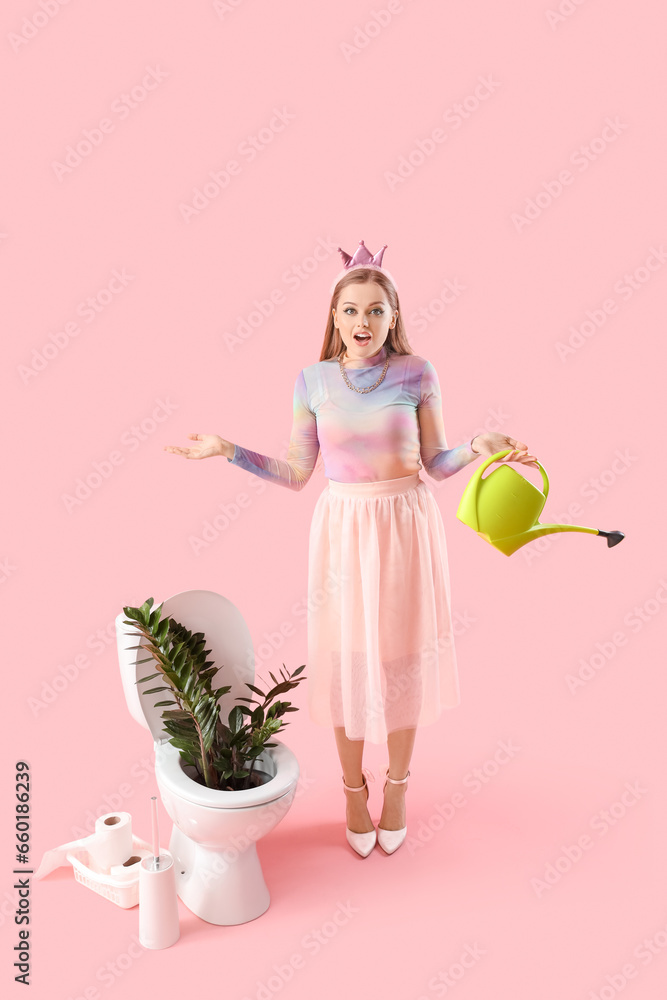 Surprised young woman with watering can and plant in toilet bowl on pink background