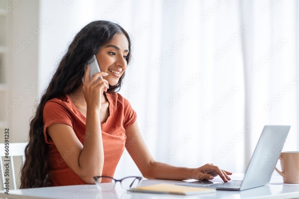 Indian lady using laptop, speaking on smartphone at home office