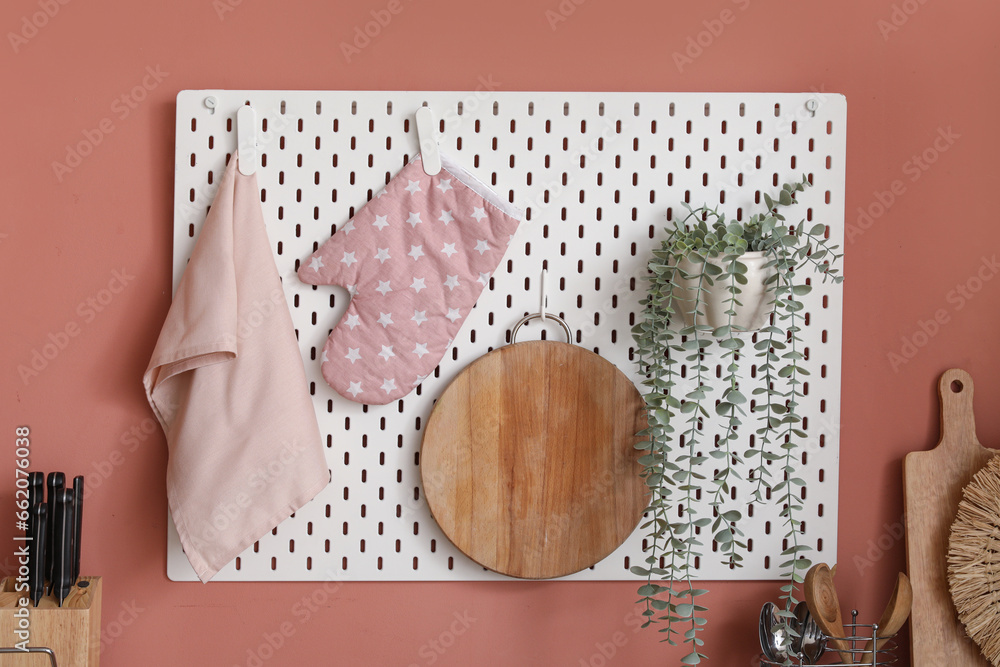 White pegboard with wooden cutting board, houseplant, mitten and towel hanging on pink wall