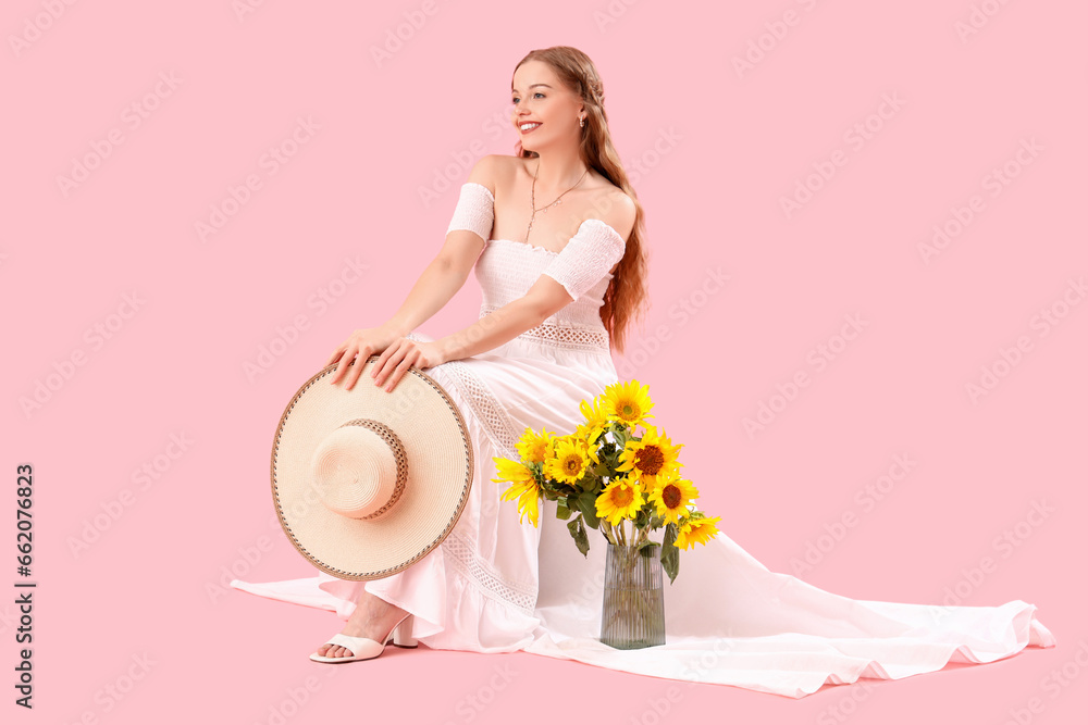 Young woman with summer hat and sunflowers in vase on pink background