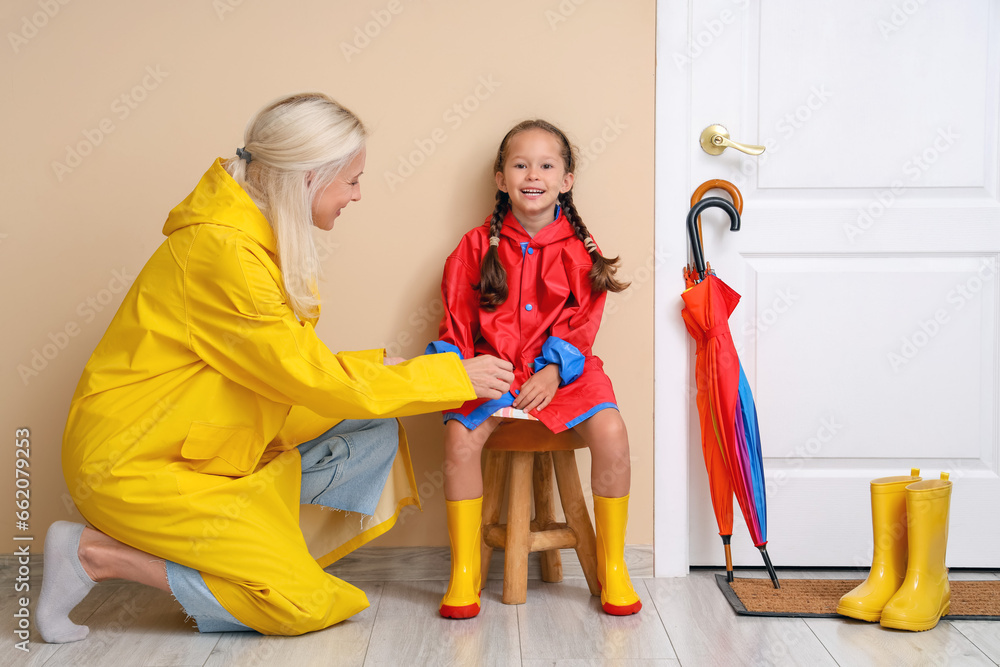 Little girl and her grandmother in raincoats getting ready for walk at home