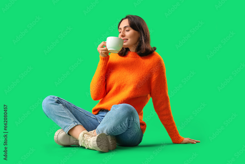 Young woman in orange sweater with cup of tea sitting on green background