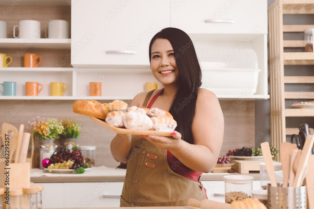 Cheerful, beautiful, chubby Asian woman stands smiling happily at bread and donut kitchen counter.