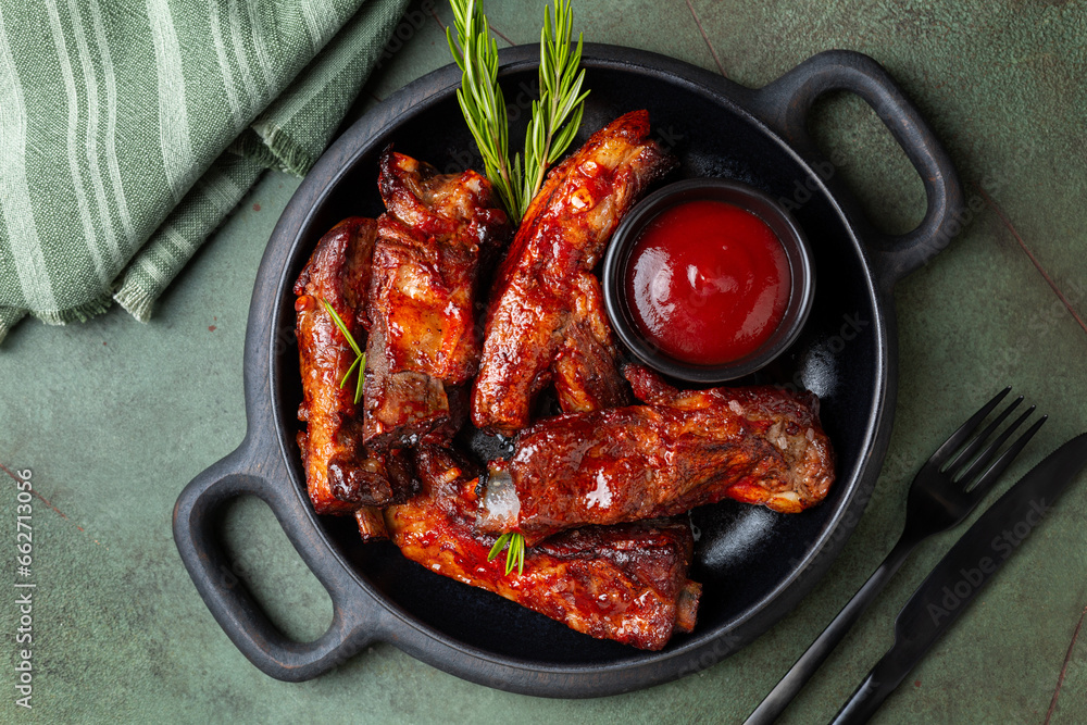 Black wooden board with baked pork ribs with barbecue sauce, rosemary. Green table background, top view. Meat food.