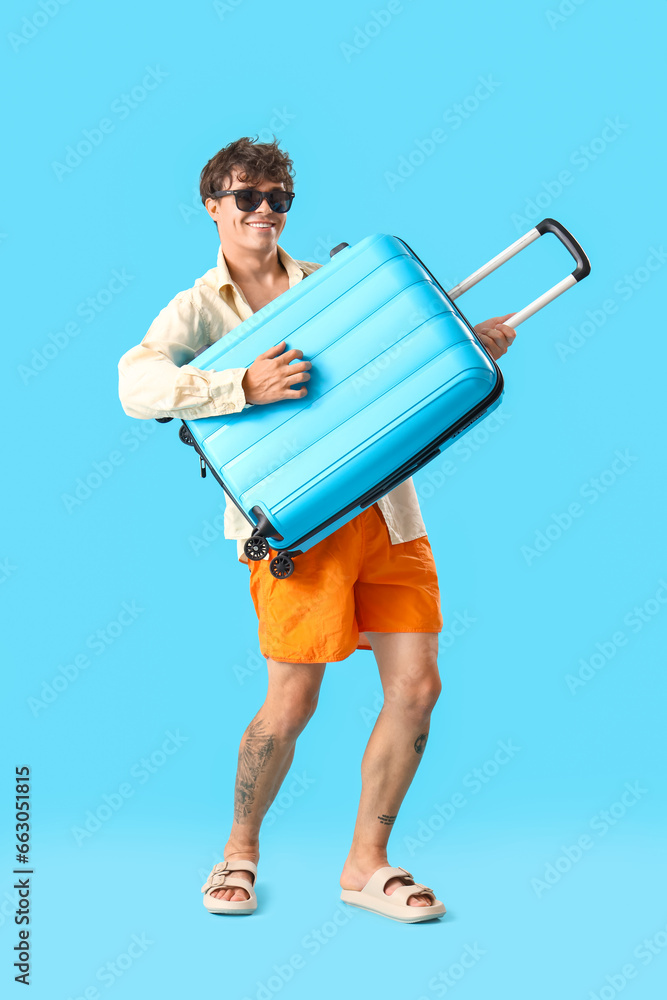 Young man in sunglasses with suitcase on blue background