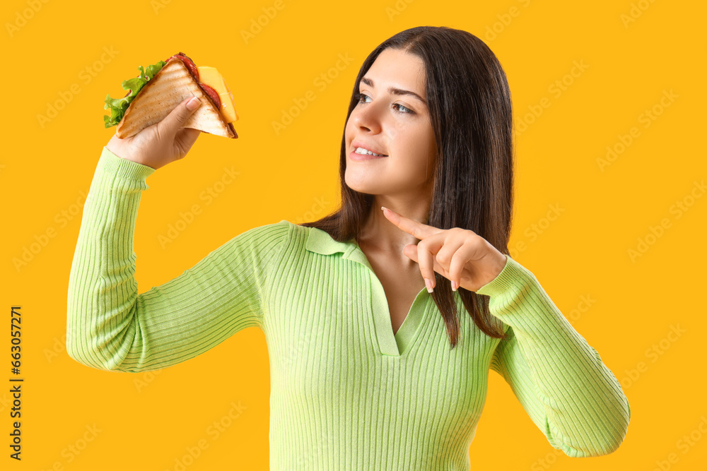 Young woman pointing at tasty sandwich on yellow background