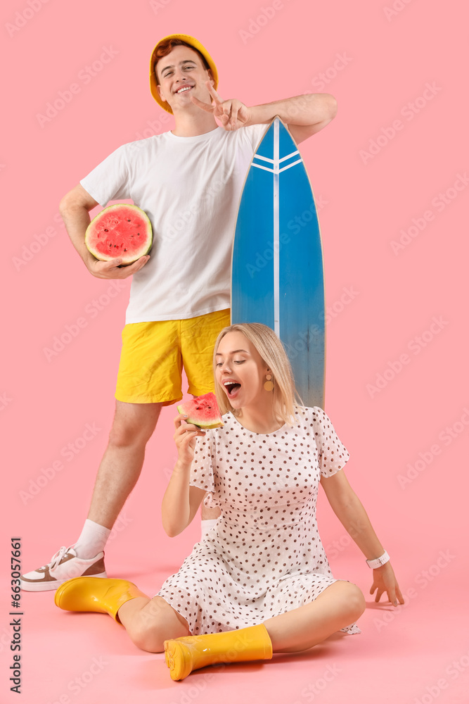 Young man with woman holding fresh watermelon and surfboard on pink background