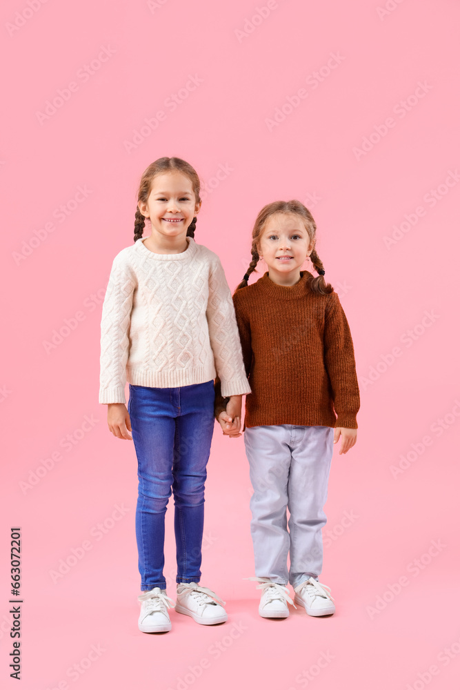 Cute little girls in warm sweaters holding hands on pink background