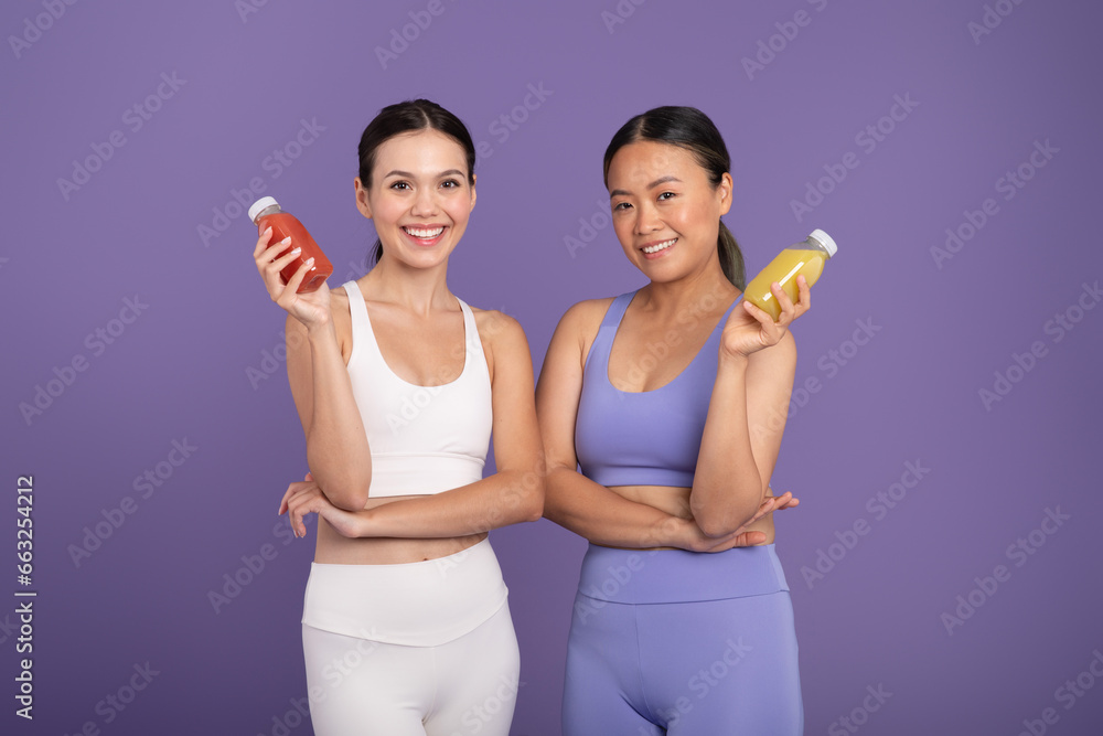 Caucasia and asian ladies enjoy smoothies post-training, posing over purple studio background