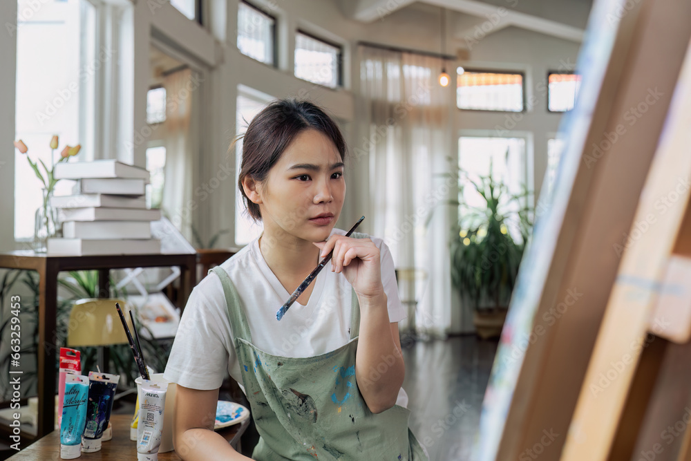 Female artist thinking and hold paintbrush while looking at canvas and pondering about paintin