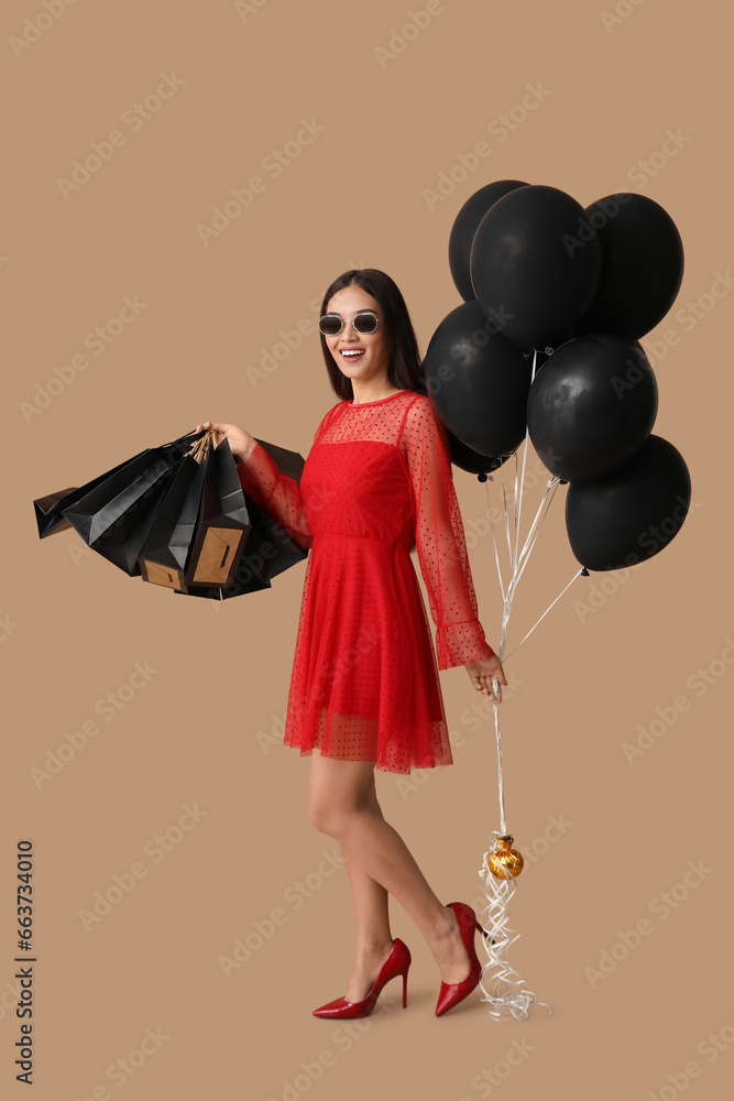 Beautiful young Asian woman with shopping bags and balloons on brown background. Black Friday sale