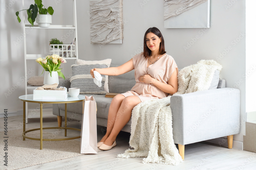 Young pregnant woman with baby clothes sitting on sofa at home