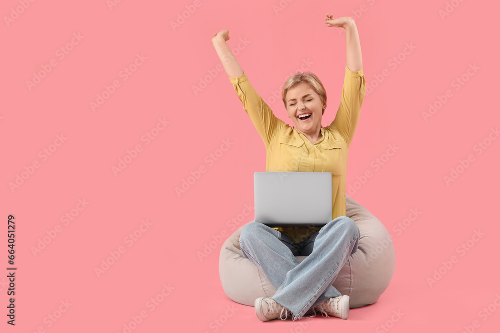 Happy young female programmer with laptop sitting on beanbag chair against pink background