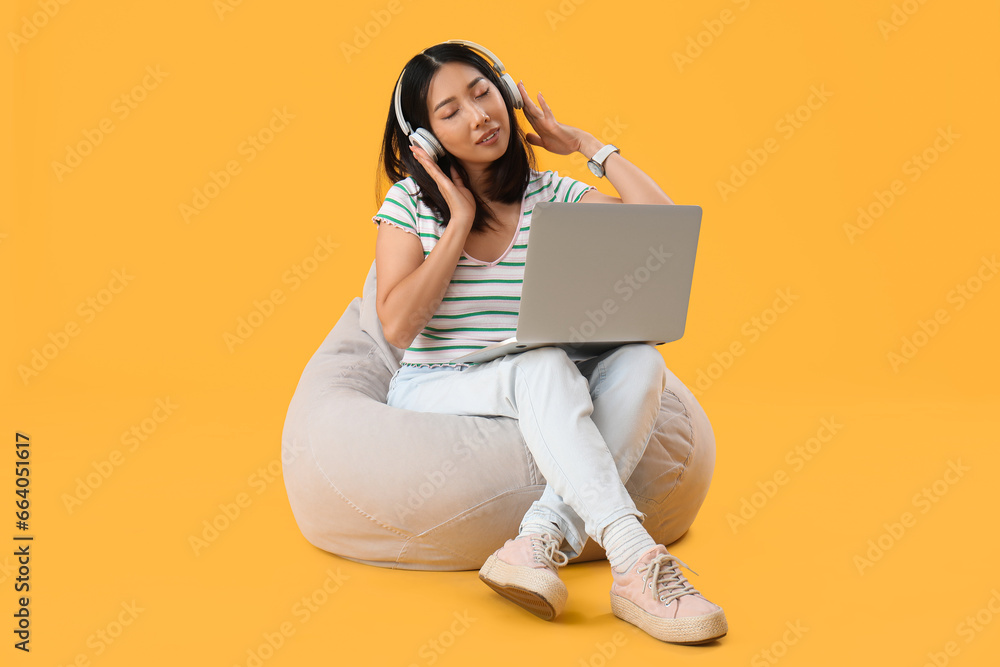 Young female Asian programmer in headphones with laptop sitting on beanbag chair against yellow background