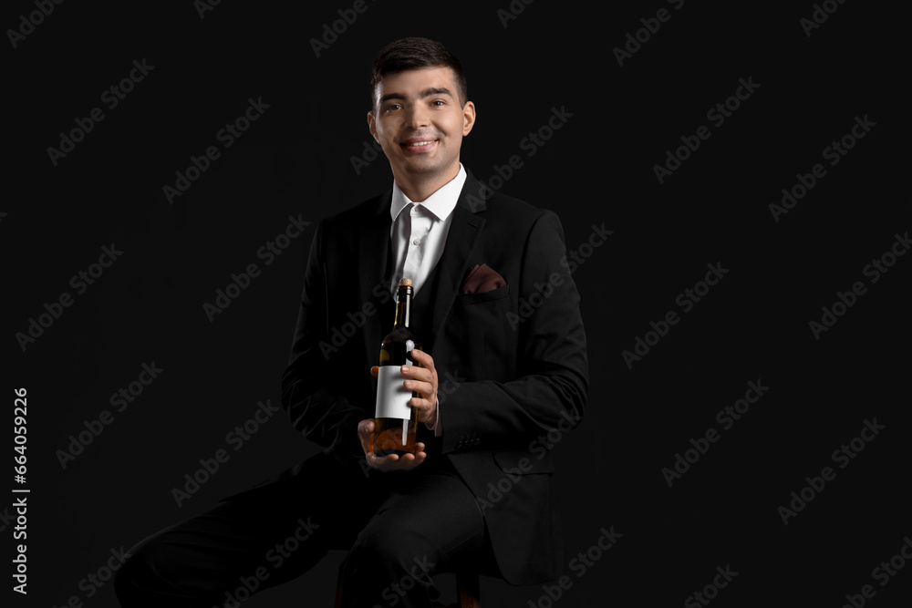 Young sommelier with bottle of wine sitting on black background