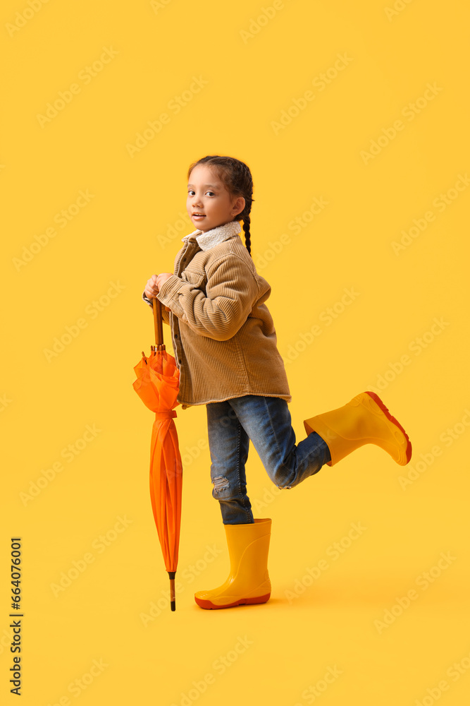 Little Asian girl in rubber boots with umbrella on yellow background