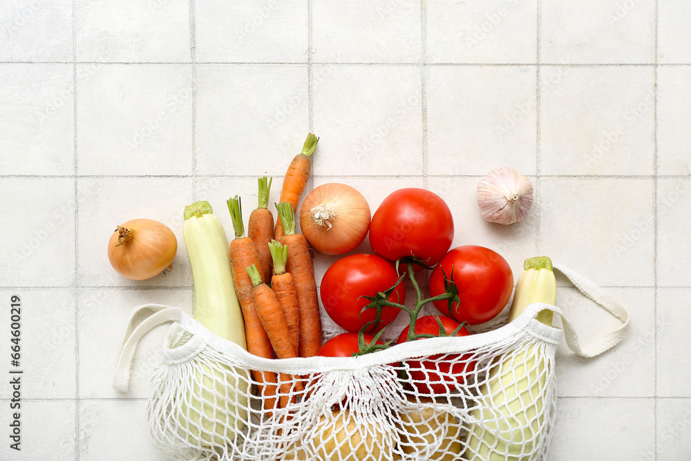 Mesh bag with different fresh vegetables on light tile background