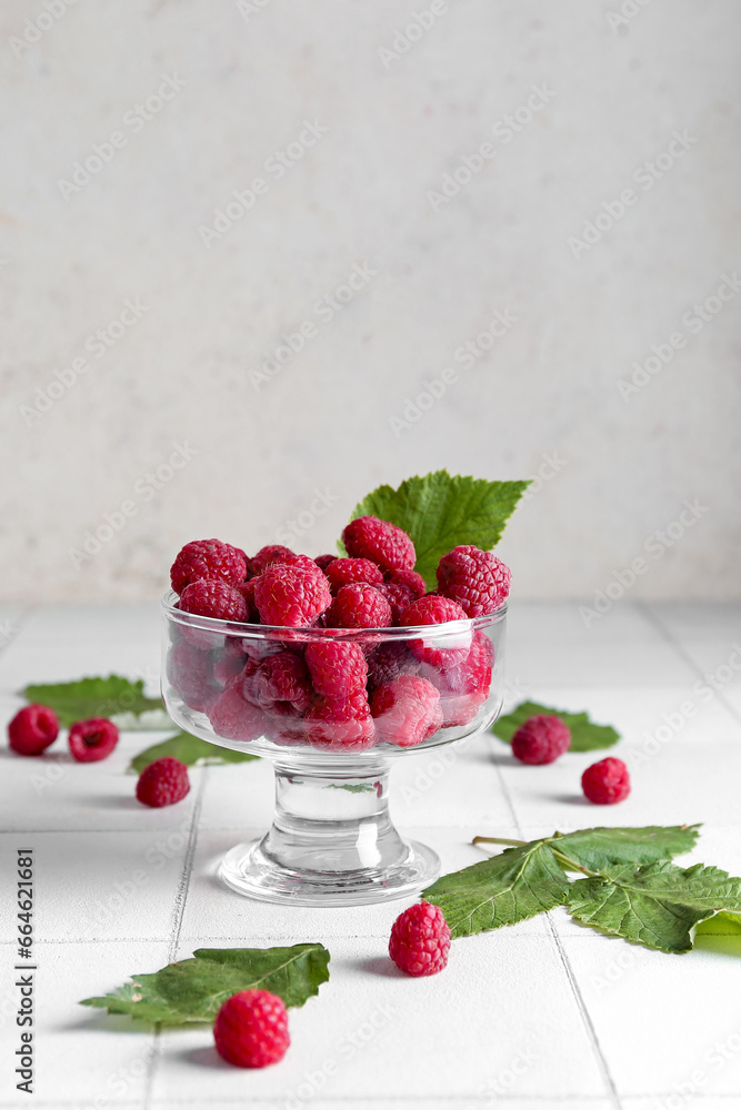 Glass bowl with fresh ripe raspberries and leaves on white tile table