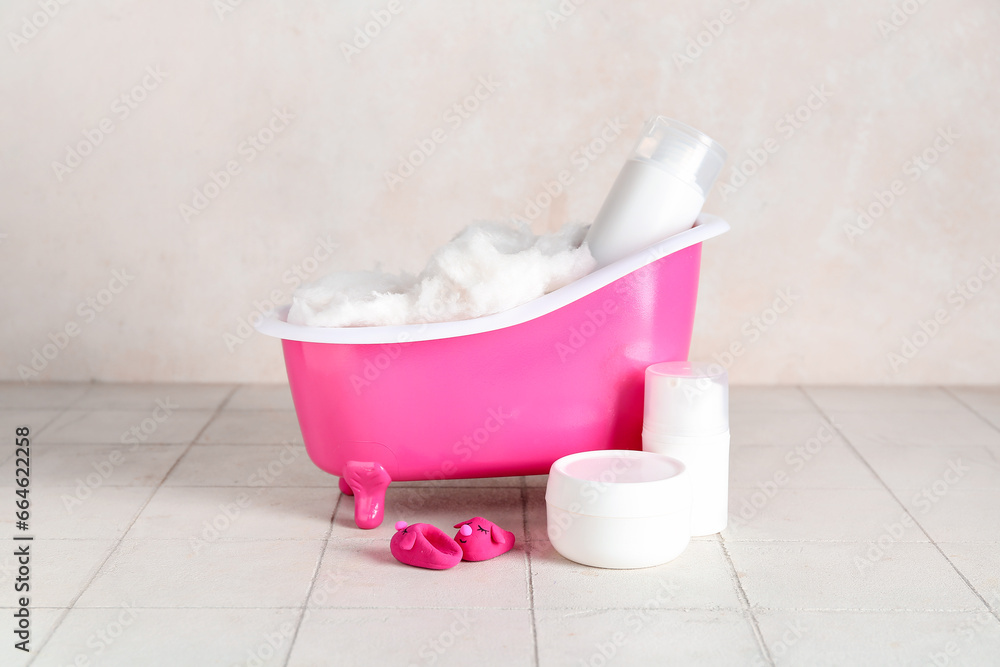 Small bathtub with cotton wool and cosmetic products on light tile table