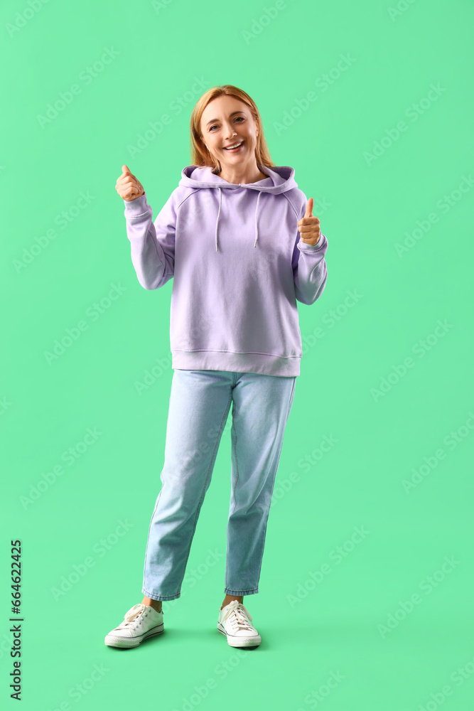 Mature woman in lilac hoodie showing thumbs-up on green background