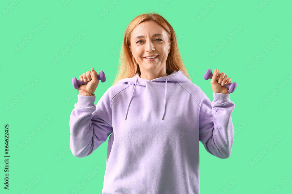 Mature woman in lilac hoodie with dumbbells on green background