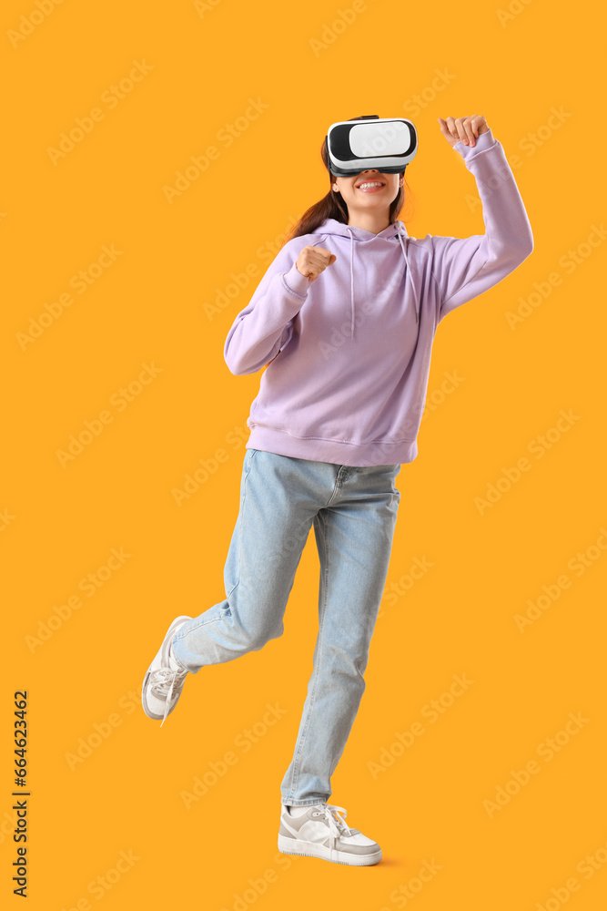 Young woman in lilac hoodie using VR glasses on yellow background