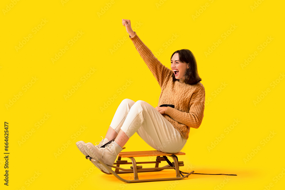 Young woman in warm sweater sitting on sledge against yellow background