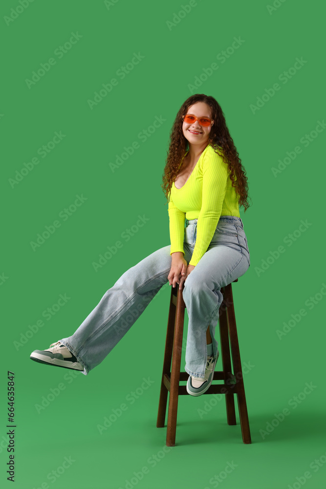 Beautiful young woman with stylish sunglasses sitting on chair against green background