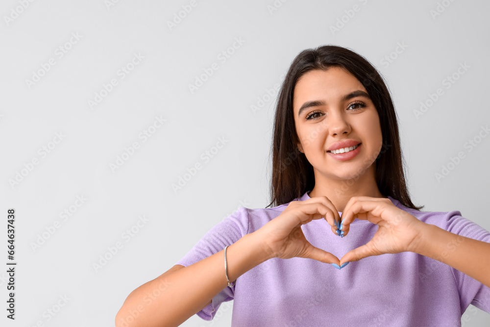 Smiling young woman with healthy teeth making heart gesture on light background