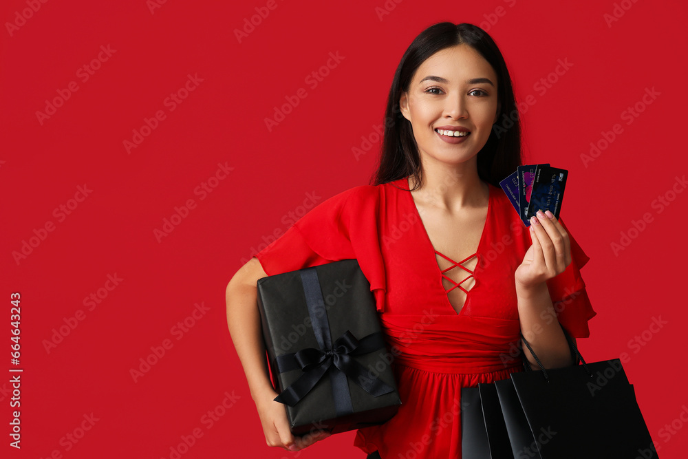 Beautiful young Asian woman with shopping bags, gift box and credit cards on red background. Black Friday sale