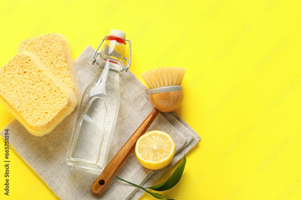 Bottle of vinegar, sponges, brush, napkin and lemon on yellow background