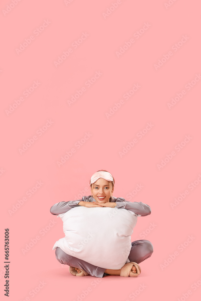 Young woman in pajamas with pillow sitting on pink background