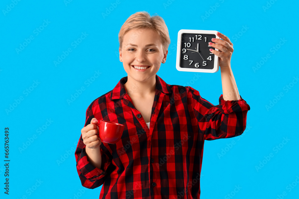 Young woman in pajamas with cup of coffee and clock on blue background