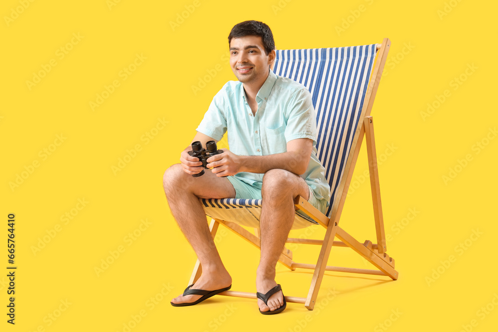 Young man with binoculars sitting in deck chair on yellow background