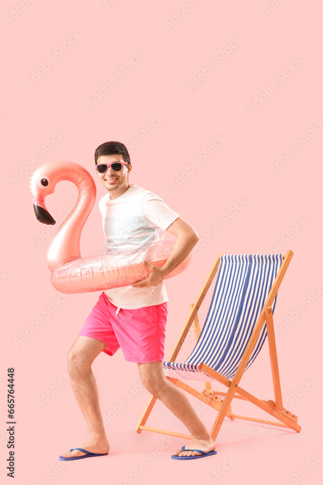 Young man with swim ring and deck chair on pink background