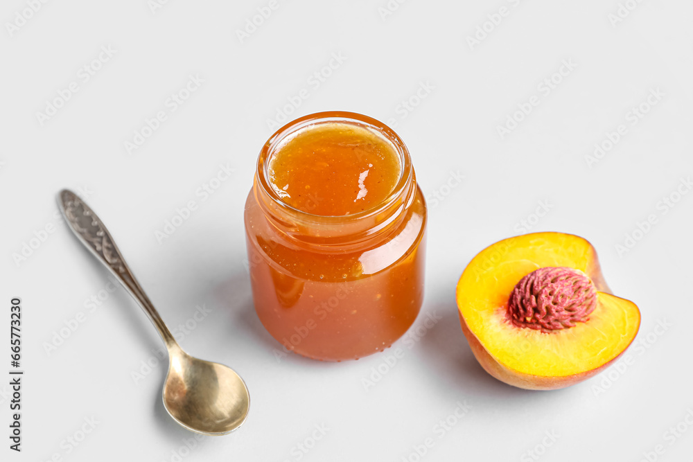 Glass jar of tasty peach jam and spoon on light background