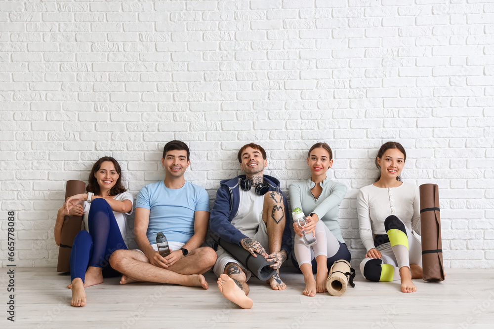 Group of sporty young people with yoga mats sitting near white brick wall