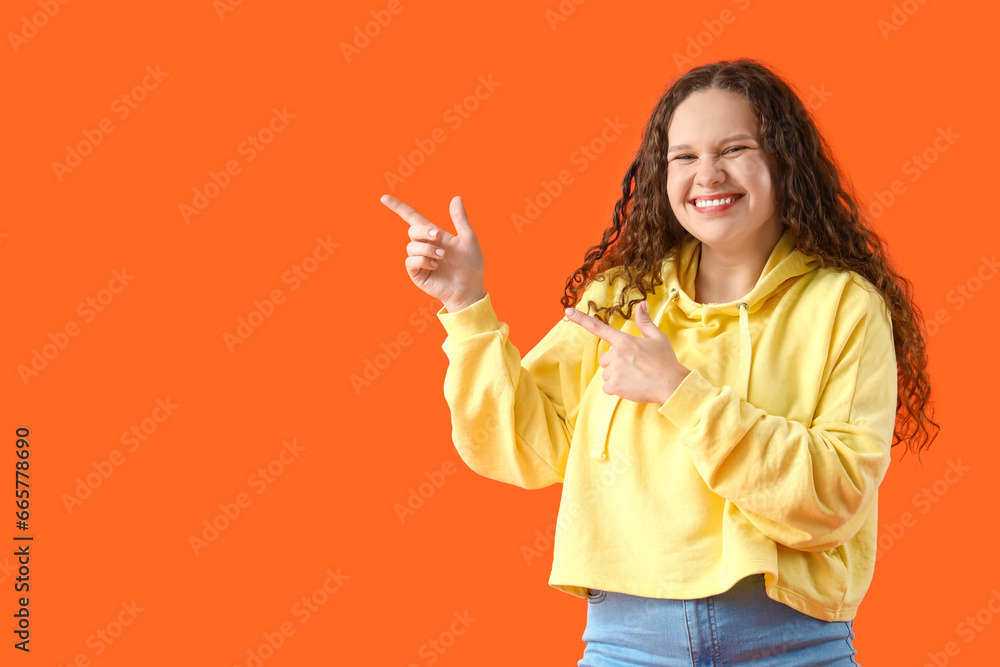 Beautiful young woman in stylish hoodie pointing at something on orange background