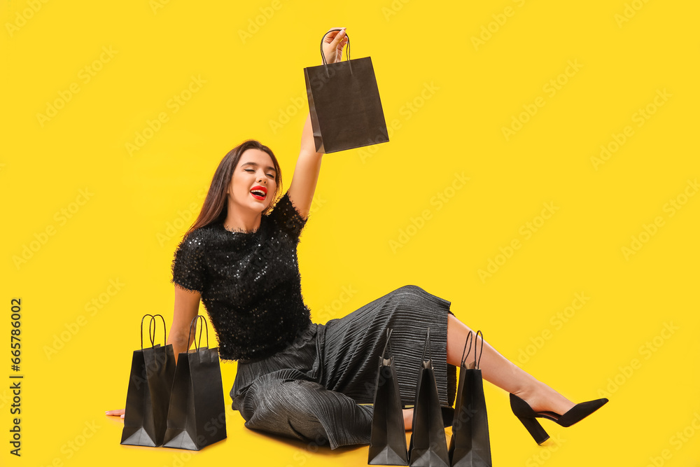 Beautiful young woman with shopping bags sitting on yellow background. Black Friday sale