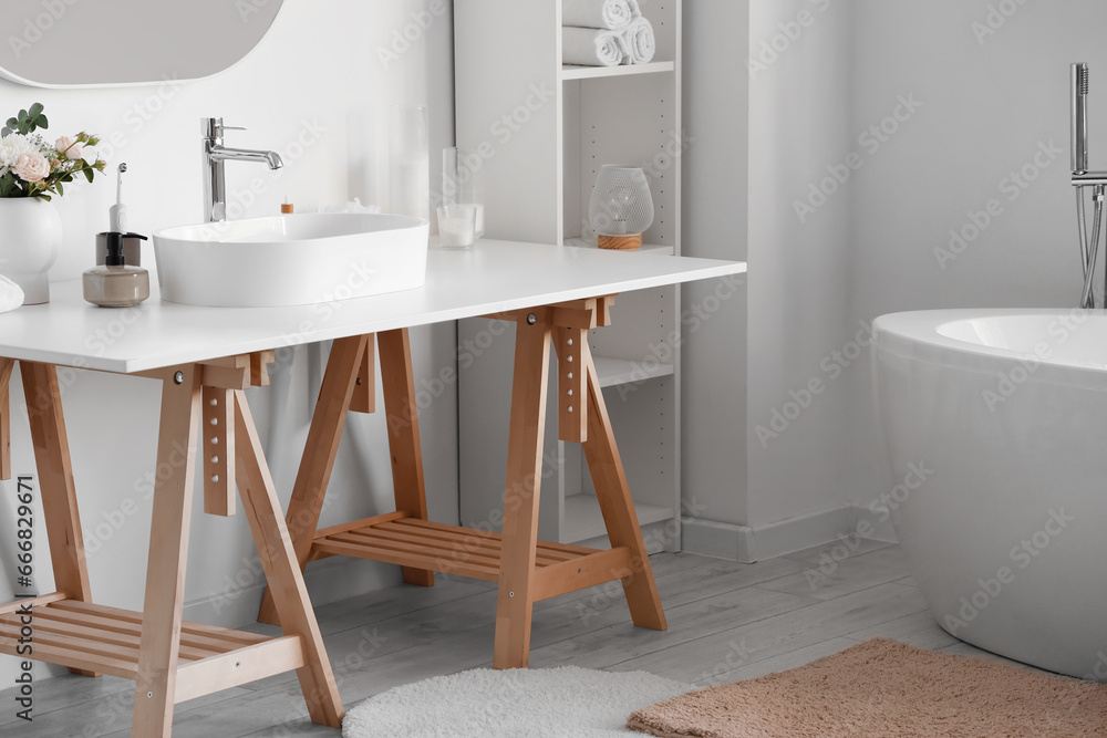 Interior of light bathroom with white sink, shelving unit and bathtub