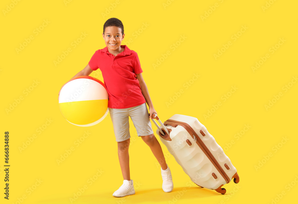 Little African-American boy with suitcase and beach ball on yellow background