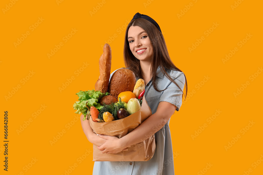 Young woman holding paper bag with fresh vegetables on yellow background