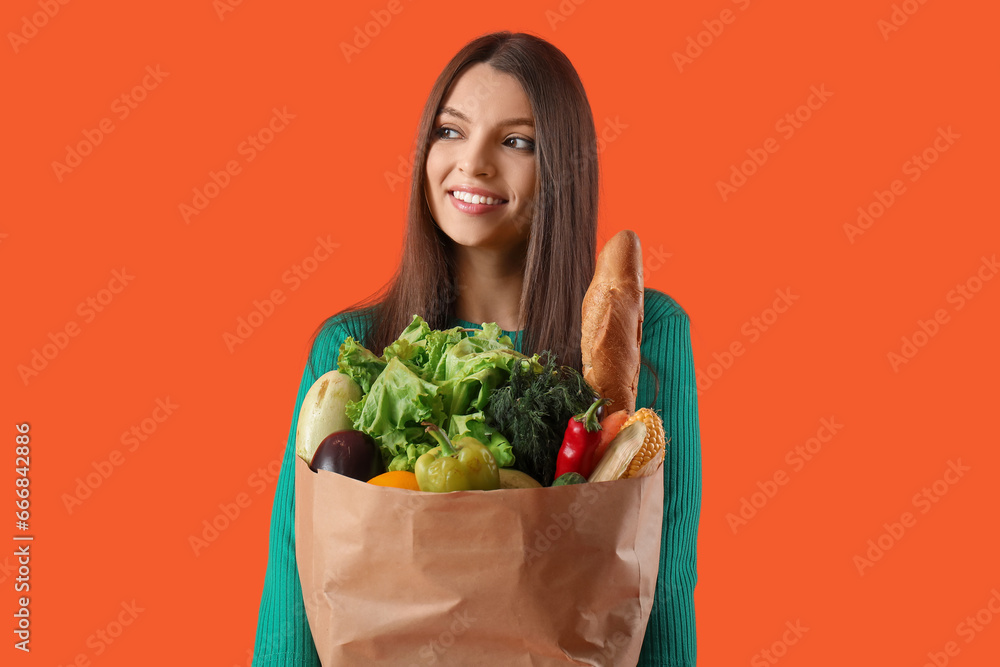 Young woman holding paper bag with fresh vegetables on orange background