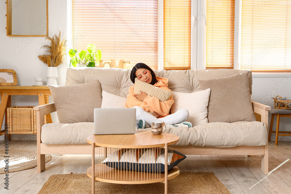 Young Asian woman in stylish hoodie with pillow sitting on sofa at home