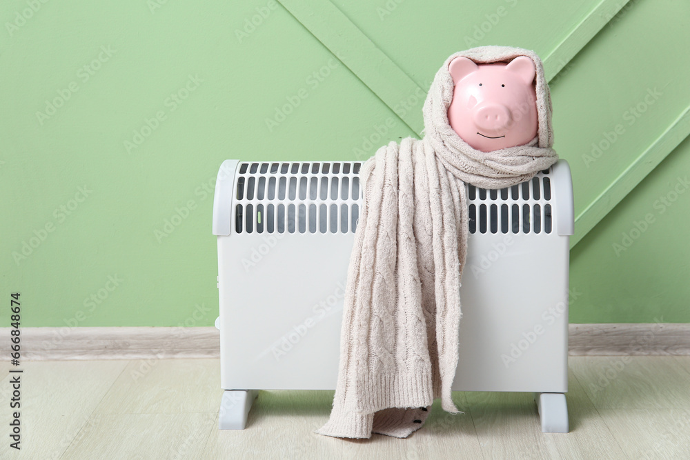 Piggy bank with warm scarf on electric convector heater near green wall at home. Heating saving concept