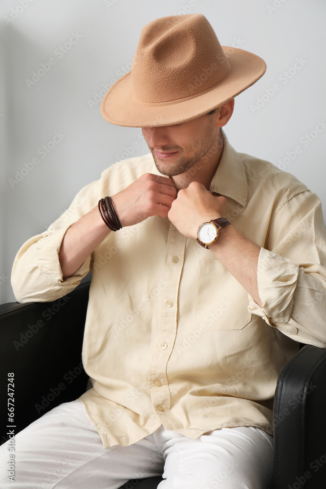 Stylish young man in hat with wristwatch adjusting shirt and sitting on armchair, closeup