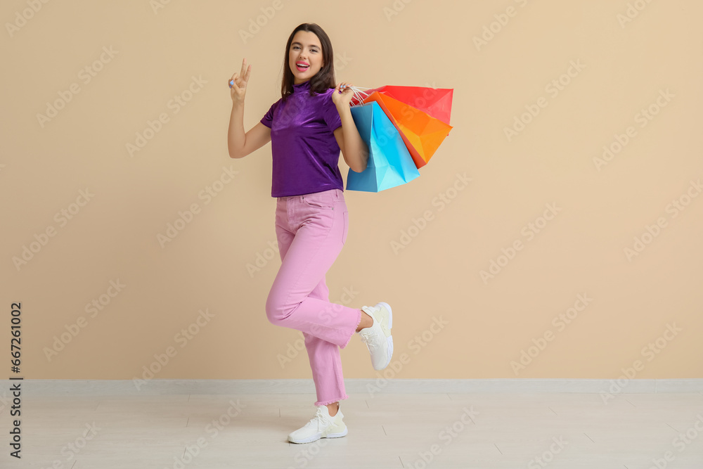 Beautiful happy young woman with shopping bags showing victory gesture near beige wall