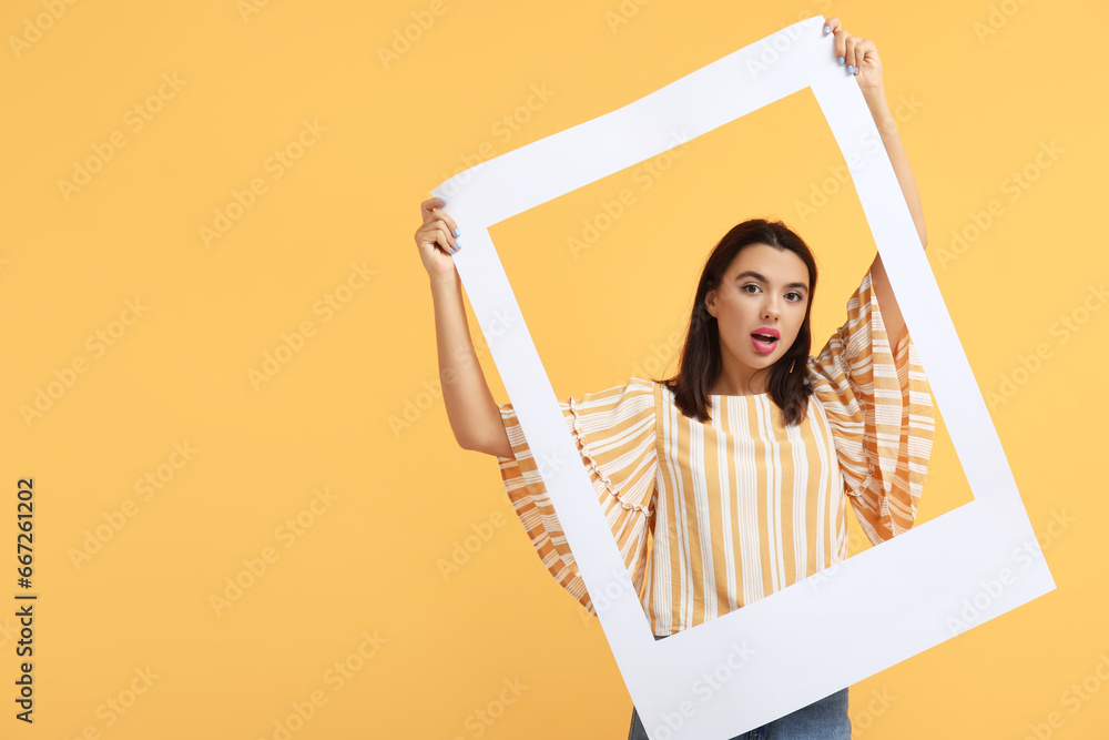 Beautiful shocked young woman with frame on yellow background