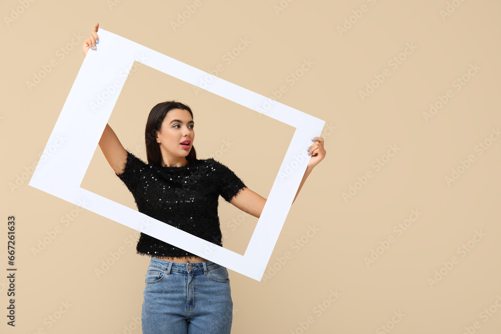 Beautiful shocked young woman with frame on beige background